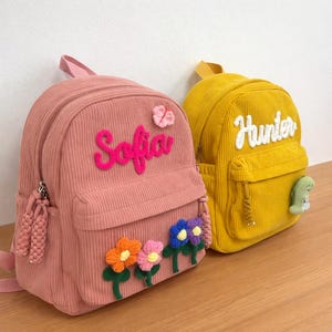 May include: Two corduroy backpacks, one pink and one yellow. The pink backpack has the name "Sofia" embroidered in pink, and flower appliques. The yellow backpack has the name "Hunter" embroidered in white, and a small dinosaur applique.