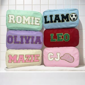 Könnte beinhalten: Eine Sammlung rechteckiger Kosmetiktaschen in verschiedenen Pastellfarben, jede mit einem Namen oder Initialen in einer Kontrastfarbe bestickt. Eine Tasche zeigt einen Fußball, eine andere einen Cowboyhut.