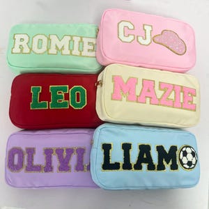 Könnte beinhalten: Sechs rechteckige Kosmetiktaschen in verschiedenen Farben, jede mit einem Namen oder Initialen in einer kontrastierenden Farbe. Die Taschen sind mintgrün, rosa, rot, cremefarben, lavendelfarben und hellblau. Einige Taschen haben zusätzliche dekorative Elemente.