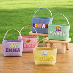 May include: Five corduroy Easter baskets in various colors, each with a handle and a name embroidered on the front. The baskets are blue, green, yellow, pink, and purple, and are sitting on a wooden surface.