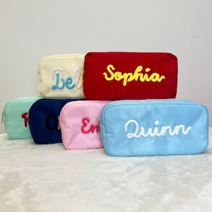 May include: A collection of six rectangular cosmetic bags in various colors, including red, blue, pink, and green. Each bag features a name embroidered in a contrasting color, such as "Sophia" in yellow on a red bag. The bags are arranged on a white surface.