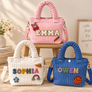 May include: Three corduroy tote bags with top handles and detachable shoulder straps. The pink bag features the name EMMA, a cherry patch, and a carrot. The white bag displays SOPHIA, a flower, hat, and rainbow. The blue bag shows OWEN and a ball.