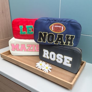 Könnte beinhalten: Vier personalisierte Kosmetiktaschen in Rot, Marineblau, Creme und Schwarz, jede mit einem Namen in einer Kontrastfarbe bestickt. Die marineblaue Tasche zeigt eine Football-Grafik. Die Taschen werden auf einem Holztablett mit zwei weißen Gänseblümchen präsentiert.