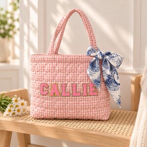 May include: A quilted tote bag featuring a pink and white gingham pattern with two top handles. The name CALLIE is displayed in pink varsity-style letters with gold trim. A blue and white patterned silk-like scarf is tied to the right handle.