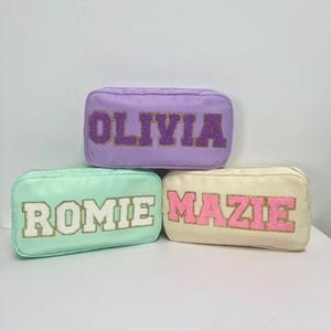 Könnte beinhalten: Drei rechteckige Kosmetiktaschen in Pastellfarben: Lavendel, Mintgrün und Creme. Jede Tasche hat einen Namen in einer kontrastierenden Farbe mit glitzernder, strukturierter Optik. Die Namen lauten "OLIVIA", "ROMIE" und "MAZIE".