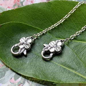 May include: A silver chain with two flower-shaped clasps. The clasps are detailed with a five-petal flower and a leaf.