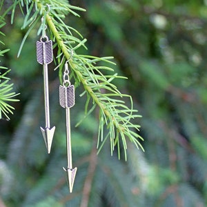 May include: A pair of silver arrow earrings hanging from a pine tree branch. The arrows have a simple design with a pointed tip and a fletching at the back.