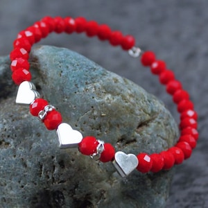 May include: Red beaded bracelet with silver heart charms. The bracelet is made of small, round, red beads and has five silver heart charms.