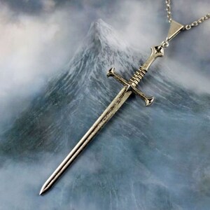 K&ouml;nnte beinhalten: Eine silberne Schwertpendantkette mit einem detaillierten Design. Das Schwert hat einen Kreuzschutz und eine spitze Spitze. Die Kette h&auml;ngt vor dem Hintergrund einer Bergkette mit Wolken.