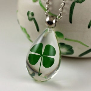May include: A clear, teardrop-shaped pendant necklace with a silver chain. The pendant encases a vibrant green four-leaf clover. The background is a white surface and a decorative ceramic pot with green leaf designs.