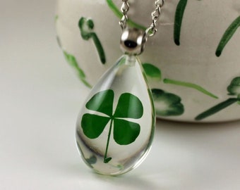 Puede incluir: Un collar con colgante transparente en forma de lágrima con una cadena plateada. El colgante encierra un trébol de cuatro hojas verde vibrante. El fondo es una superficie blanca y una maceta de cerámica decorativa con diseños de hojas verdes.
