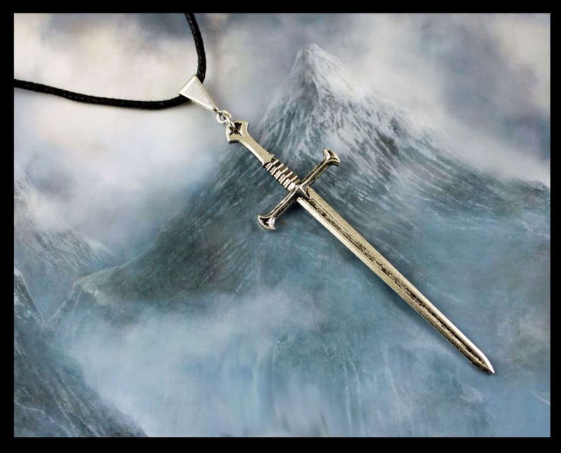 K&ouml;nnte beinhalten: Eine silberne Schwertpendantkette mit einem schwarzen Band. Das Schwert hat einen Kreuzschutz und eine spitze Spitze. Der Anh&auml;nger h&auml;ngt vor dem Hintergrund einer Bergkette mit Wolken.