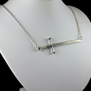May include: A silver chain necklace with a silver sword pendant. The sword is pointed and has a detailed design.