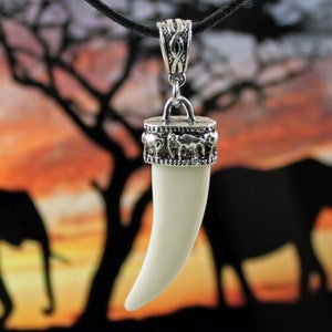 May include: A white faux ivory tusk pendant with a silver metal top featuring a design of elephants. The pendant is hanging from a black cord.