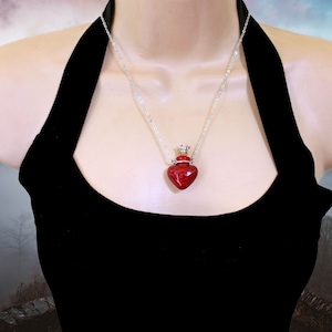 May include: A silver chain necklace with a red heart-shaped glass bottle pendant. The bottle has a silver crown-shaped stopper.
