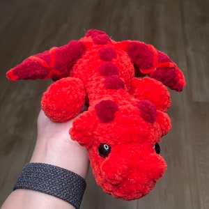 May include: A handmade, plush dragon toy in shades of red and orange. The dragon has a textured, fuzzy body with darker red accents on its back and head. It features large, black eyes and is designed to be soft and cuddly.