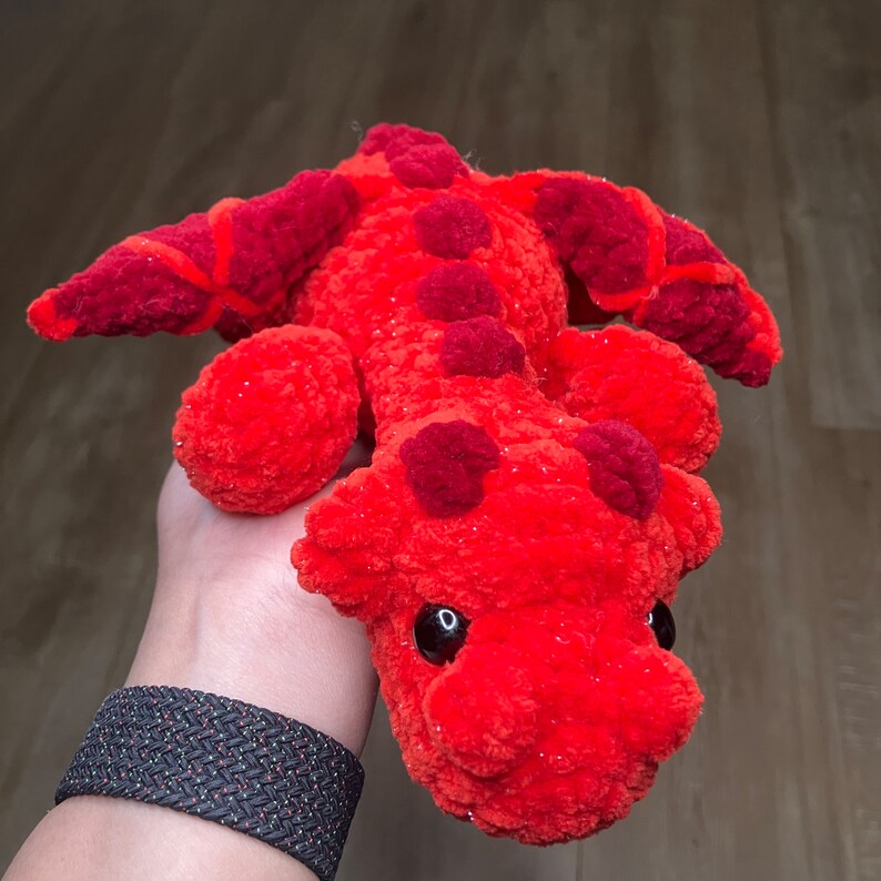 May include: A handmade, plush dragon toy in shades of red and orange. The dragon has a textured, fuzzy body with darker red accents on its back and head. It features large, black eyes and is designed to be soft and cuddly.