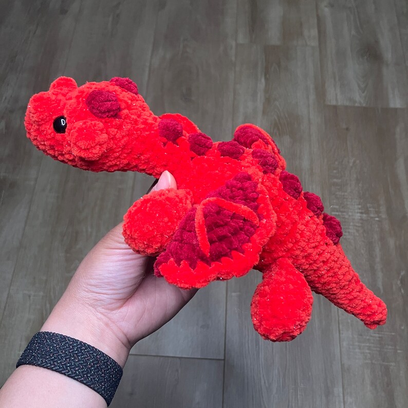 May include: A handmade, plush dinosaur toy in shades of red and burgundy. The crocheted dinosaur features a textured body, small black eyes, and darker red spots along its back. The wings and belly are a deeper shade of red.