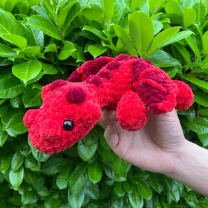 May include: A handmade red crochet dinosaur toy with dark red spots and accents. The plush toy has black eyes and is held against a backdrop of green leaves. The dinosaur is made of soft, textured yarn.