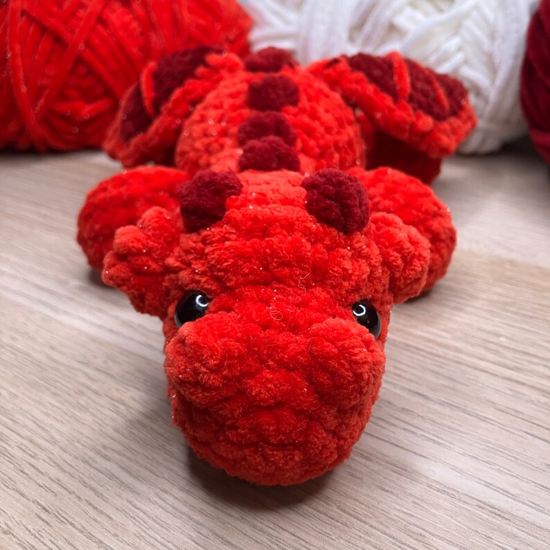May include: A handmade, plush red dragon toy with dark red accents. The dragon has large, black eyes and is made of a soft, textured yarn. The dragon is lying down, with its head facing the viewer.