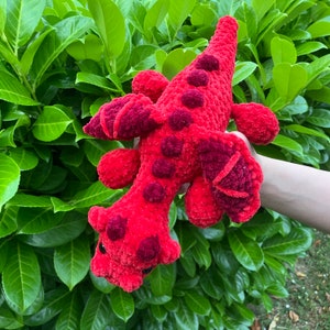 May include: A handmade, red crochet dinosaur plush toy with darker red spots and accents. The soft, textured toy features a long tail, small legs, and a detailed head. The plush is set against a backdrop of green foliage.