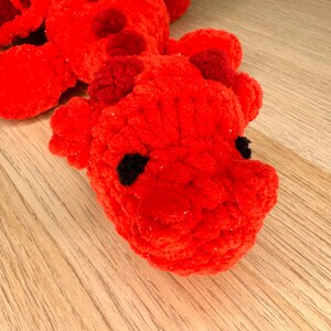 May include: A handmade, red crocheted dragon toy with black eyes and small horns. The dragon is made of a soft, textured yarn and is lying on a wooden surface. The toy is a vibrant shade of red.
