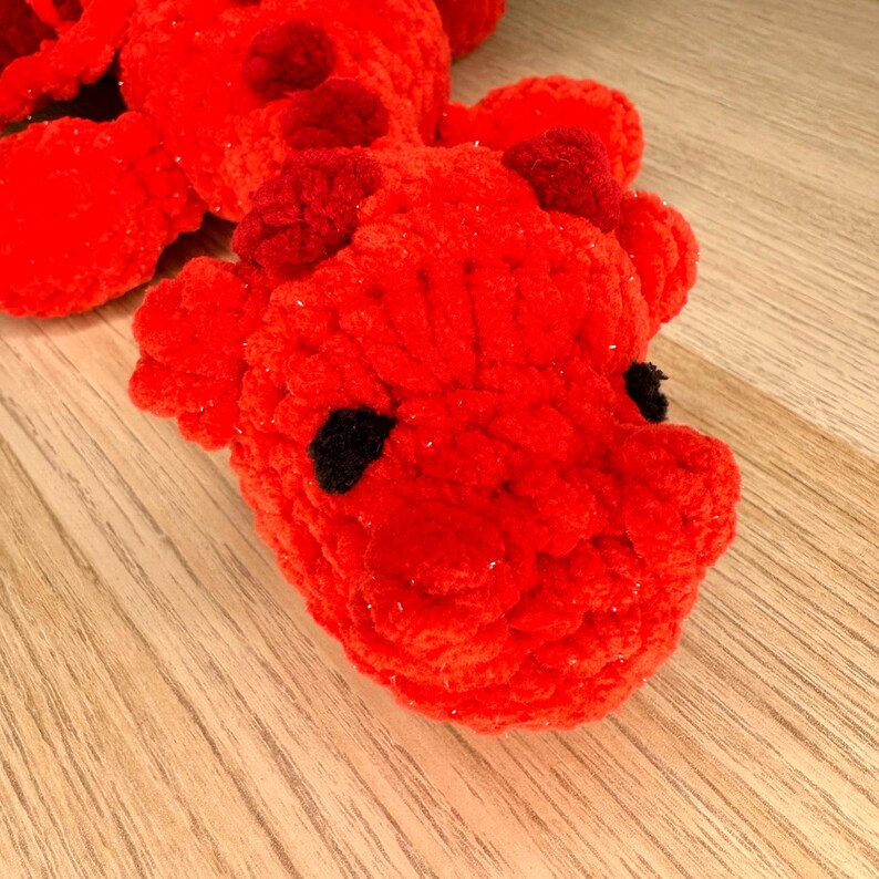 May include: A handmade, red crocheted dragon toy with black eyes and small horns. The dragon is made of a soft, textured yarn and is lying on a wooden surface. The toy is a vibrant shade of red.