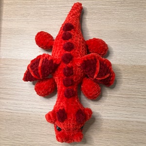 May include: A handmade, plush red alligator toy with darker red accents. The alligator has small black eyes and is made of a soft, textured material. The toy features a long body, four small legs, and a tail.