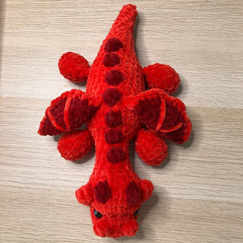 May include: A handmade, plush red alligator toy with darker red accents. The alligator has small black eyes and is made of a soft, textured material. The toy features a long body, four small legs, and a tail.