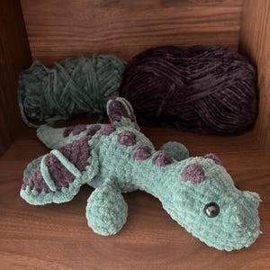 May include: A handmade, plush sea serpent toy in shades of teal and purple. The toy features a textured, velvety appearance with purple spots and accents. Two balls of yarn in teal and purple are in the background.