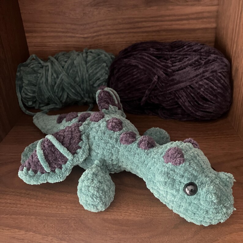 May include: A handmade, plush sea serpent toy in shades of teal and purple. The toy features a textured, velvety appearance with purple spots and accents. Two balls of yarn in teal and purple are in the background.