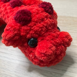 May include: Close-up of a handmade, plush red creature with dark red accents and black button eyes. The soft, textured toy has a whimsical design, perfect for a child's room or as a unique gift.