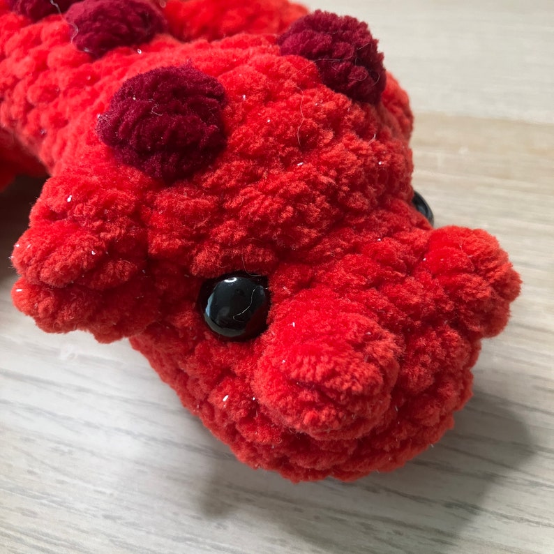May include: Close-up of a handmade, plush red creature with dark red accents and black button eyes. The soft, textured toy has a whimsical design, perfect for a child's room or as a unique gift.