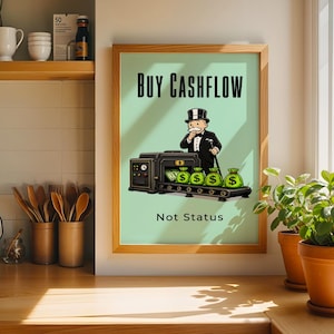 Könnte beinhalten: Ein gerahmter Druck mit dem Text "BUY CASHFLOW" über einer Illustration einer Monopoly-ähnlichen Figur und Geldsäcken. Der Text "Not Status" steht darunter. Der Rahmen ist holzfarben und der Hintergrund hellgrün.