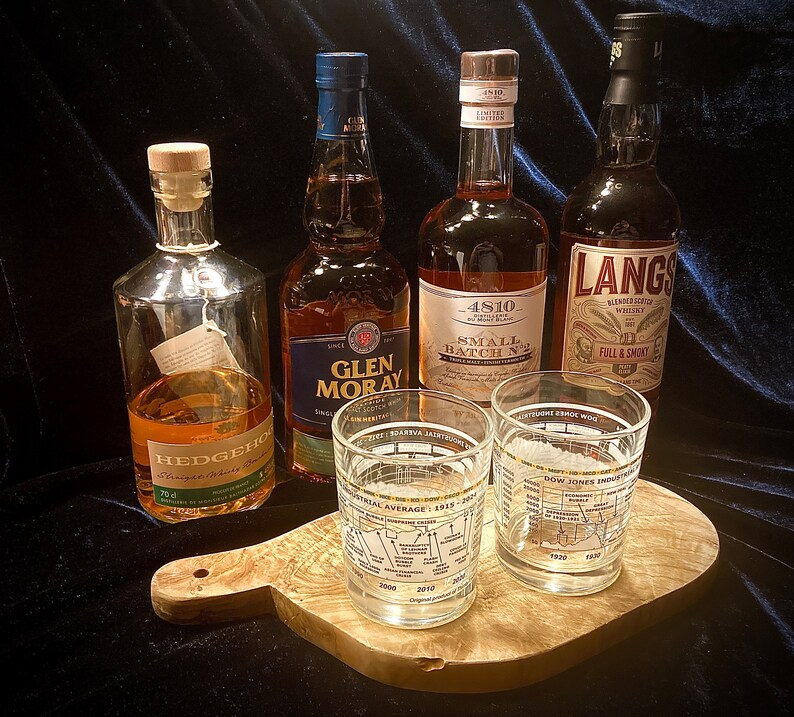 May include: An assortment of liquor bottles and two glasses on a wooden cutting board. The bottles include brands like Glen Moray, 4810, and Langs. The glasses feature a chart design with text. The bottles are various sizes and shapes.