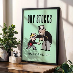 Op de afbeelding: Een ingelijst print met een groene achtergrond waarop een cartoonillustratie te zien is van Monopoly Man die een muntje geeft aan een kleiner cartoonfiguur. De tekst op de print luidt "KOOP AANDELEN, GEEN SNOEP".