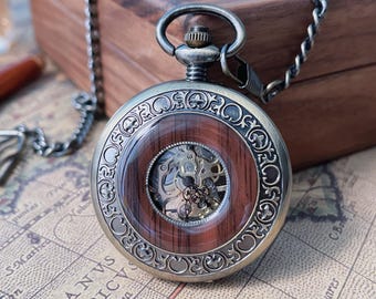 May include: Antique pocket watch with a silver-tone case and a clear face revealing the intricate mechanical movement. The watch features a dark wood-grain center and decorative silver-tone detailing around the face. A silver chain is attached.