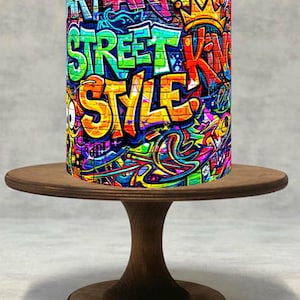 Könnte beinhalten: Ein zylindrisches Objekt mit lebendiger Graffiti-Kunst, das die Wörter "ART", "STREET KING STYLE" und ein Kronen-Design zeigt. Es steht auf einem kleinen, runden Kuchenständer aus Holz und präsentiert einen kühnen, farbenfrohen urbanen Stil.