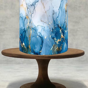 Pode incluir: Um objeto cilíndrico com um design abstrato azul e dourado, semelhante a mármore ou arte em tinta, está sobre um suporte de bolo de madeira. O design apresenta tons de azul com detalhes dourados. O suporte é feito de madeira escura.