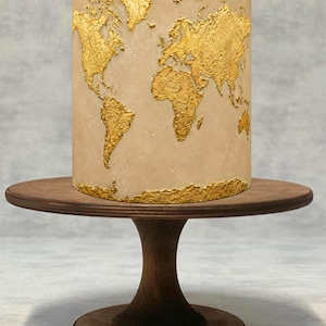 Puede incluir: Un pastel cilíndrico con un acabado liso en color beige claro que presenta un mapa del mundo texturizado en color oro metálico alrededor de su exterior. El pastel está colocado sobre un soporte de madera marrón oscuro con base circular.