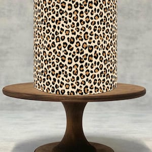 Può includere: Un paralume cilindrico con un motivo leopardato, con macchie nere su sfondo beige. Il paralume è posto su un alzatina per torte in legno marrone scuro con piano rotondo e base a piedistallo. Il supporto è in legno.