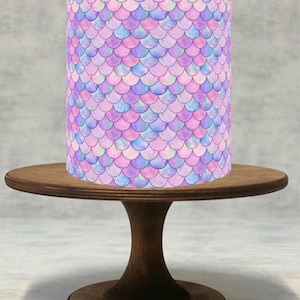 May include: A cylindrical object with a mermaid scale pattern in shades of pink, purple, and blue sits on a small, round, wooden cake stand. The stand has a single, curved pedestal base.
