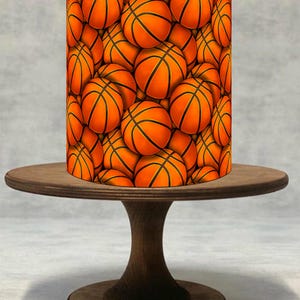 May include: A cylindrical lampshade with a repeating pattern of orange basketballs and black lines. It is displayed on a dark brown wooden cake stand with a round top and a pedestal base. The background is a light grey.