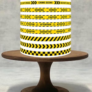 Puede incluir: Un pastel cilíndrico con diseño de cinta de advertencia, con texto como "DO NOT ENTER" y "CRIME SCENE". El pastel está sobre un soporte de pastel de madera oscura. El diseño incluye rayas y chevrones amarillos y negros.