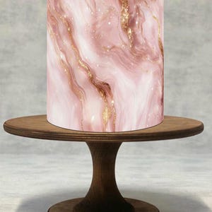 Pode incluir: Um suporte de bolo cilíndrico com um design de mármore rosa e detalhes dourados está sobre um suporte de bolo de madeira. O suporte de bolo tem um fundo rosa claro e branco com veias douradas.