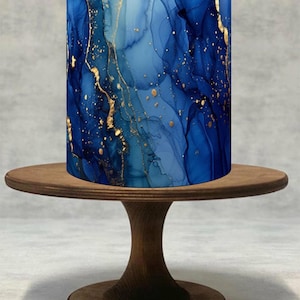 Può includere: Una torta cilindrica con un design astratto blu e oro è posta su un alzatina di legno. Il design della torta presenta sfumature di blu con accenti dorati, creando un effetto marmorizzato. L'alzatina è in legno scuro.