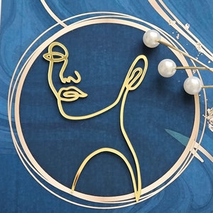 Puede incluir: Obra de arte en metal dorado que representa un rostro minimalista dentro de un círculo dorado, sobre un fondo azul oscuro veteado. Tres cuentas blancas con forma de perla están unidas a finos alambres dorados. La obra tiene una estética abstracta y moderna.