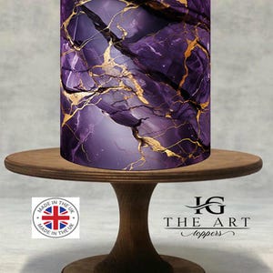 May include: A cylindrical cake topper with a purple and gold marble design, displayed on a wooden cake stand. The topper features a swirling pattern of deep purple and gold veins. The cake stand is made of dark wood.