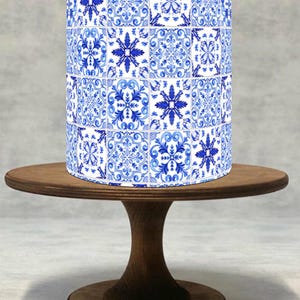 Könnte beinhalten: Ein zylindrisches Objekt mit einem blau-weißen Fliesenmuster, das an traditionelle portugiesische Azulejo-Fliesen erinnert, steht auf einem kleinen, runden Kuchenständer aus Holz. Der Ständer hat eine dunkelbraune Oberfläche und einen Sockel.