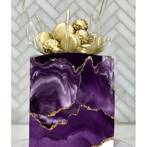 May include: A decorative cake with a purple and gold marbled design. The cake is topped with gold-colored spheres and a clear, abstract glass structure. The cake sits on a white cake stand.
